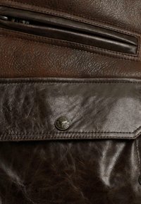 Bruine leren tas met een gestructureerde oppervlakte, bovenste ritssluiting en een opvallende metalen knoopaccent. Gedetailleerde stiksels en een zichtbare zak.