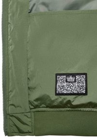 Žalia striukė su lygiu, blizgiu paviršiumi ir matiniais rankogaliais. Yra juodas etiketė su QR kodu ir logotipu. Detalus vaizdas iš vidaus.
