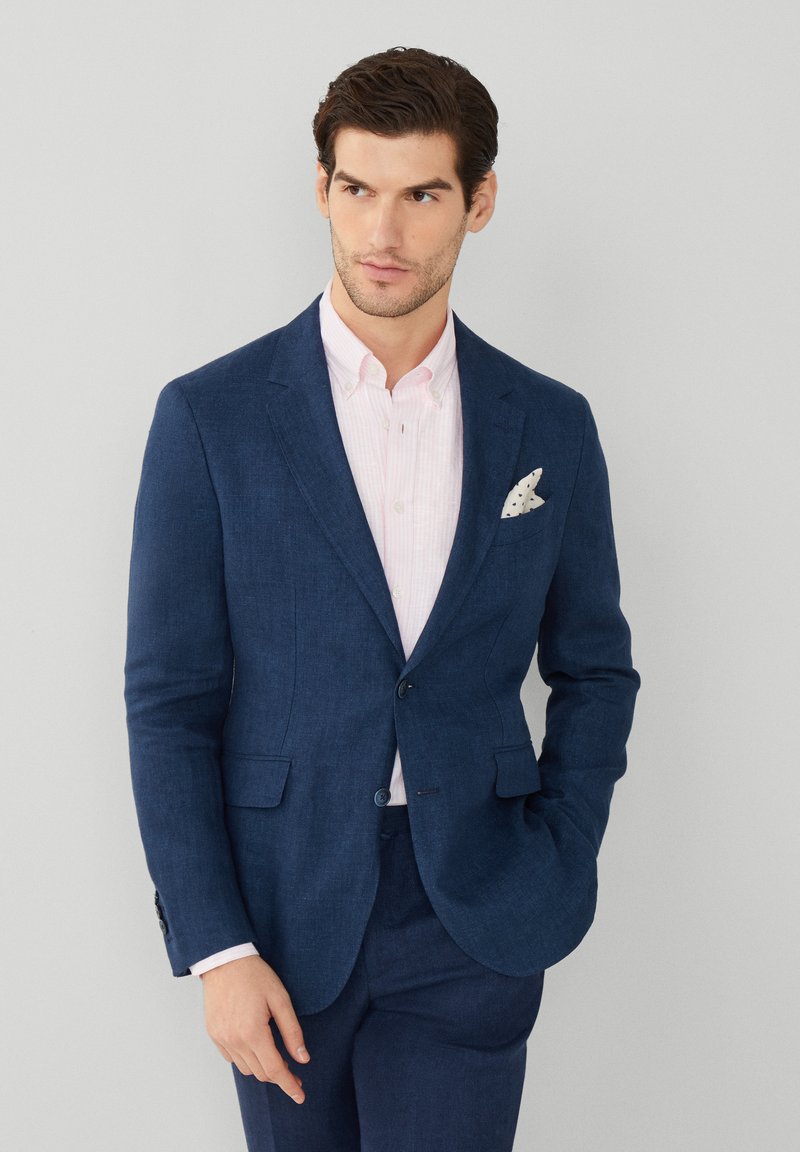 Mörkblå blazer med en texturerad yta, i kombination med en ljusrosa randig skjorta och en vit bröstnäsduk med svarta prickar.
