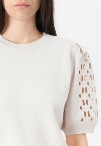 Lichtbeige trui met een ronde hals en textuurpuffmouwen, voorzien van opengewerkte geometrische patronen aan de zijkanten. Zacht materiaal.