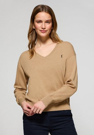 Beżowy sweter w serek wykonany z miękkiego materiału, z długimi rękawami, ściągaczami oraz małym znakiem po lewej stronie.