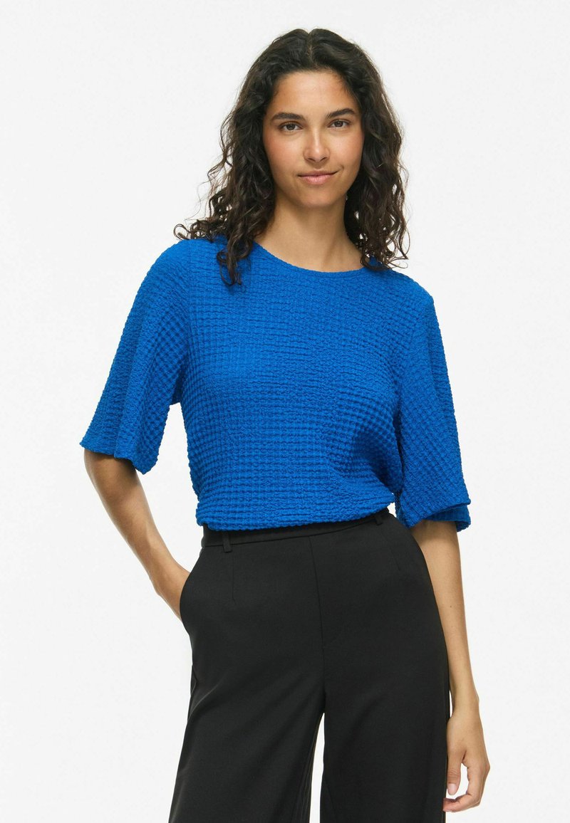 Femme aux cheveux bouclés et foncés porte un haut texturé bleu vif à manches courtes et un pantalon noir taille haute, debout sur un fond blanc.