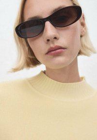 Gafas de sol negras en forma ovalada sobre un suéter amarillo claro de canalé con un cuello alto. Las gafas tienen lentes ahumados y un diseño elegante.