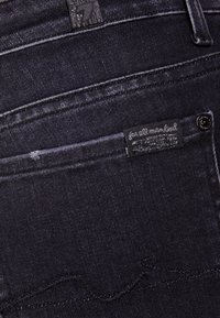 Tejanos de denim oscuro con un diseño ajustado, que presentan un ligero desgaste, costuras negras y una etiqueta con el texto "para toda la humanidad".