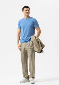 Blaues Rundhals-T-Shirt, beige Hose und weiße Turnschuhe. Das Model hält eine beige Jacke. Glattes Material, lockerer Schnitt und lässiger Stil.