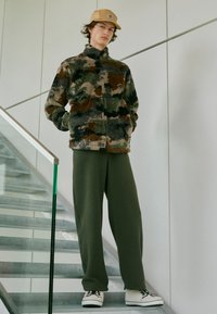 Fleecejakke med camouflage mønster i grøn, brun og sort. Sammen med olivengrønne fleecebukser og hvide sneakers med sorte detaljer.