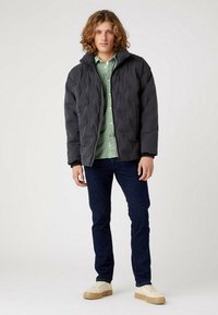 Chaqueta negra acolchada con cuello alto, con un diseño acolchado. Llevada sobre una camisa verde de botones y combinada con jeans oscuros y zapatos claros.