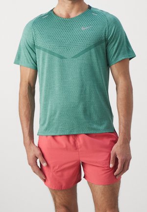 Mand iført grøn mønstret kortærmet sportstrøje og koralrøde sportsshorts står mod hvid baggrund.