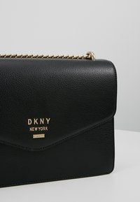 Svart läderhandväska med en strukturerad yta, med en vikt flik och en kedjerem i guldton. Märkesnamnet är präglat i guld på framsidan.