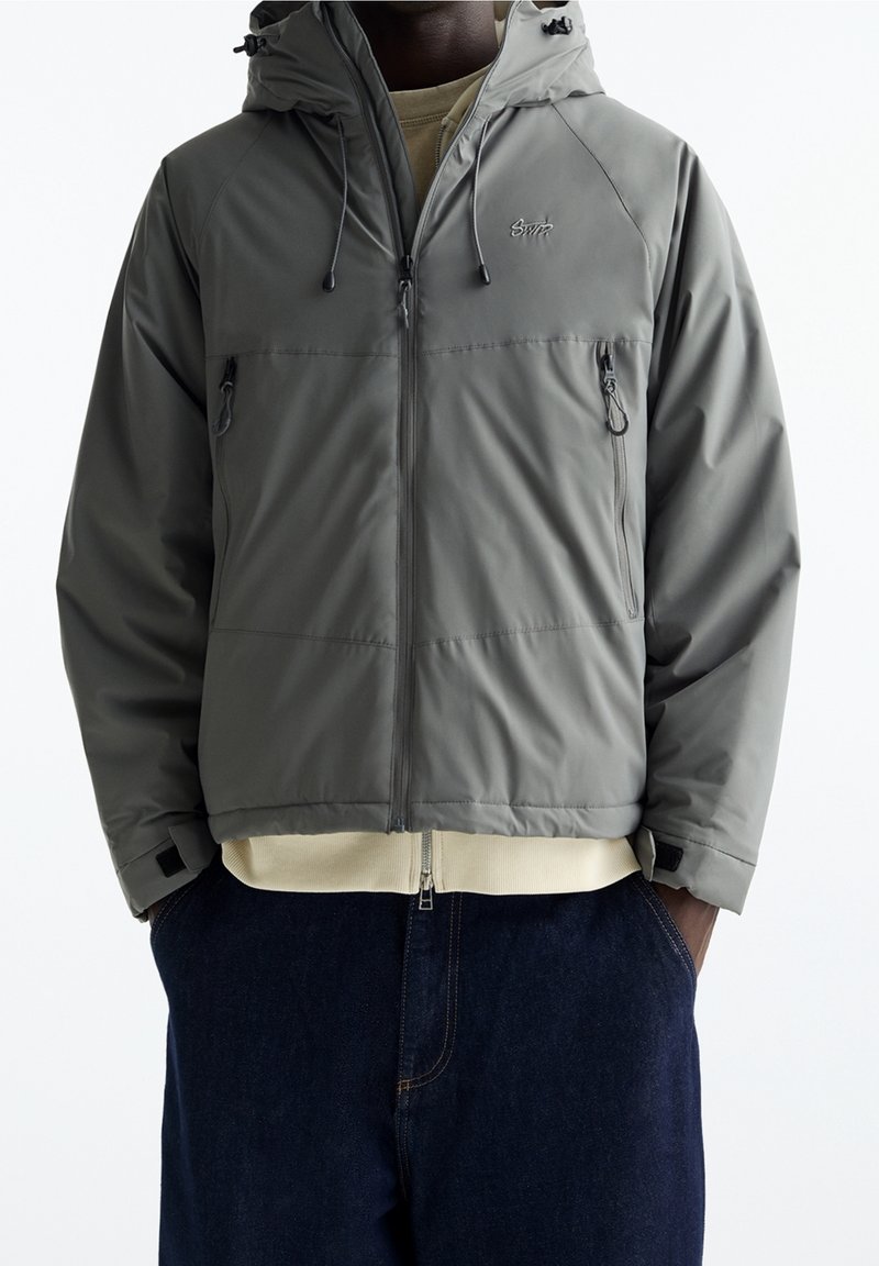 Homme portant une veste à capuche grise zippée avec deux poches poitrine zippées et un pantalon bleu foncé, les mains dans les poches.