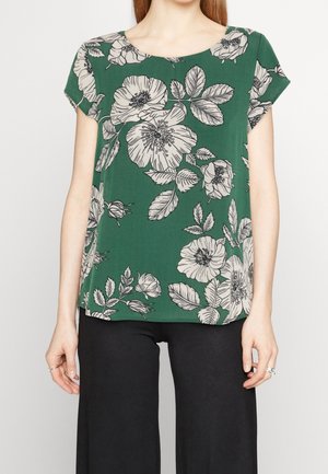 Groene korte mouw blouse met groot wit bloemmotief, gedragen met zwarte broek, toont bovenlichaam en armen.