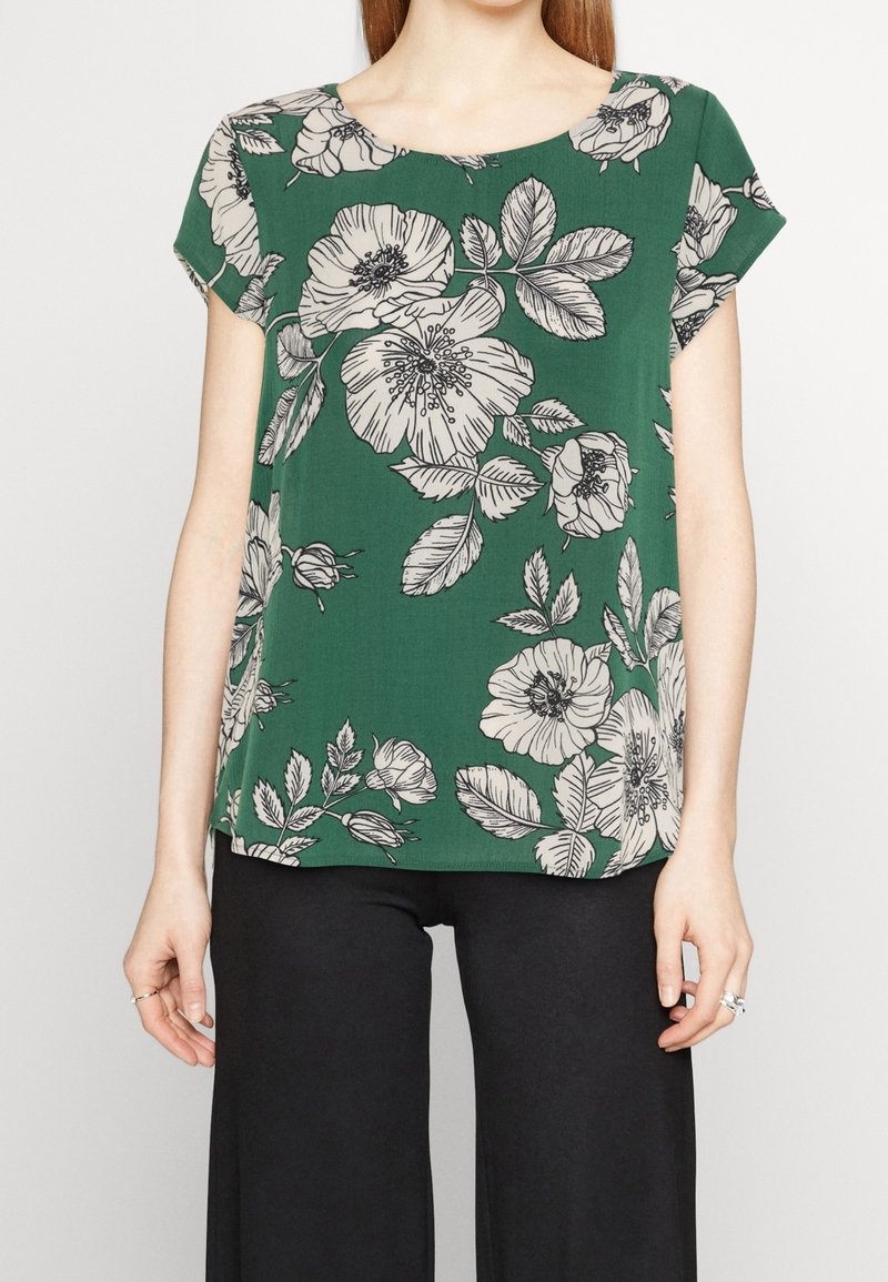 Blouse verte à manches courtes avec un grand imprimé floral blanc portée avec un pantalon noir, montrant le haut du torse et les bras.