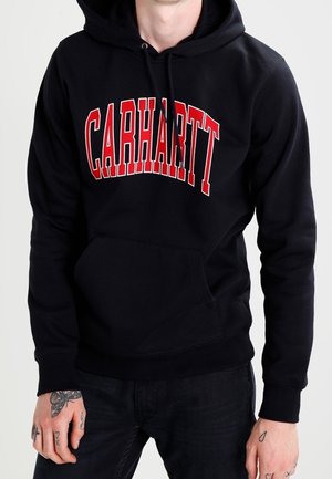 Osoba nosząca czarną bluzę z kapturem z czerwonym napisem „CARHARTT”, w połączeniu z czarnymi spodniami, pokazująca wytatuowane dłonie.