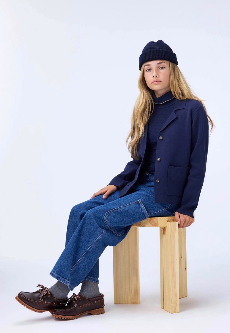 Blazer texturizado azul marino con botones plateados, combinado con un cuello de tortuga azul marino, jeans de pierna ancha azules y zapatos marrones con cordones rosas, sentado en un taburete de madera.