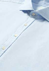 Camicia azzurra chiaro con bottoni, realizzata in un tessuto morbido. Presenta un colletto, sei bottoni e una superficie leggermente testurizzata.