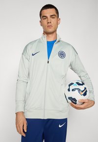 Lysegrå zip-up jakke med blåt logo, båret over en blå t-shirt. Personen holder en mønstret hvid og blå fodbold.