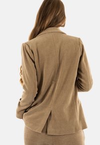 Blazer in velluto cord di colore marrone chiaro con vestibilità sartoriale, presenta spalle pieghettate e un dettaglio di spacco sul retro, visto da dietro.