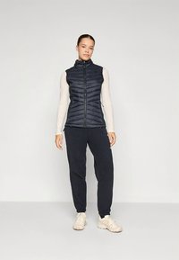 Tamsiai mėlynas pūstas liemenė su aukštu apykakliu, priekyje su užtrauktuku ir vertikaliomis siūlėmis, derinamas su juodomis siaurėjančiomis kelnėmis ir šviesios spalvos sportbačiais.