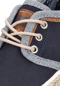 Zapatilla de lona azul marino con acentos de denim, cordones beige, ojales metálicos y parche de cuero marrón. Material texturizado con costuras visibles.