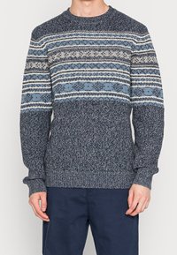 Homme portant un pull en tricot bleu et gris à motifs géométriques et floraux, associé à un pantalon bleu foncé.