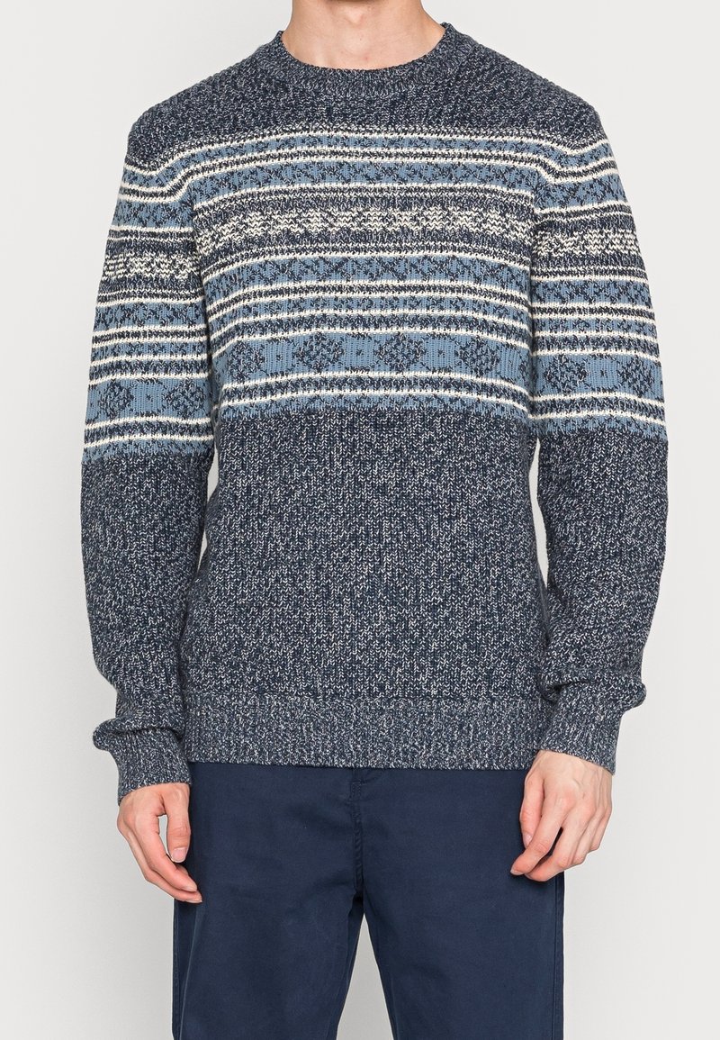 Homme portant un pull en tricot bleu et gris à motifs géométriques et floraux, associé à un pantalon bleu foncé.
