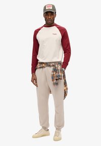 Bordeauxrood en crème raglan mouwtop, beige zwembroek, geruite shirt om de taille geknoopt, grijze pet en beige sneakers. Casual outfit met gecoördineerde kleuren.