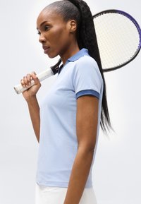 Γυναίκα τενίστρια κρατά ρακέτα στον ώμο, φοράει γαλάζιο πόλο και λευκή φούστα, δείχνει συγκεντρωμένη μπροστά σε απλό φόντο.