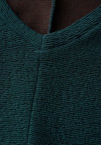 Tessuto di un teal scuro e caratterizzato da una trama a coste, con scollatura a V e una fodera interna marrone a contrasto. La superficie appare morbida e leggera.