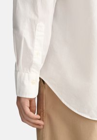 Bílá košile s dlouhým rukávem a knoflíkovým manžetami, která má hladkou texturu a špičatý design manžet, v kombinaci s khaki kalhotami.