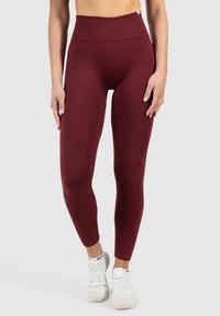Leggings bordeaux taille haute en tissu extensible, avec une texture lisse et une coupe moulante, associés à des chaussures de sport blanches.