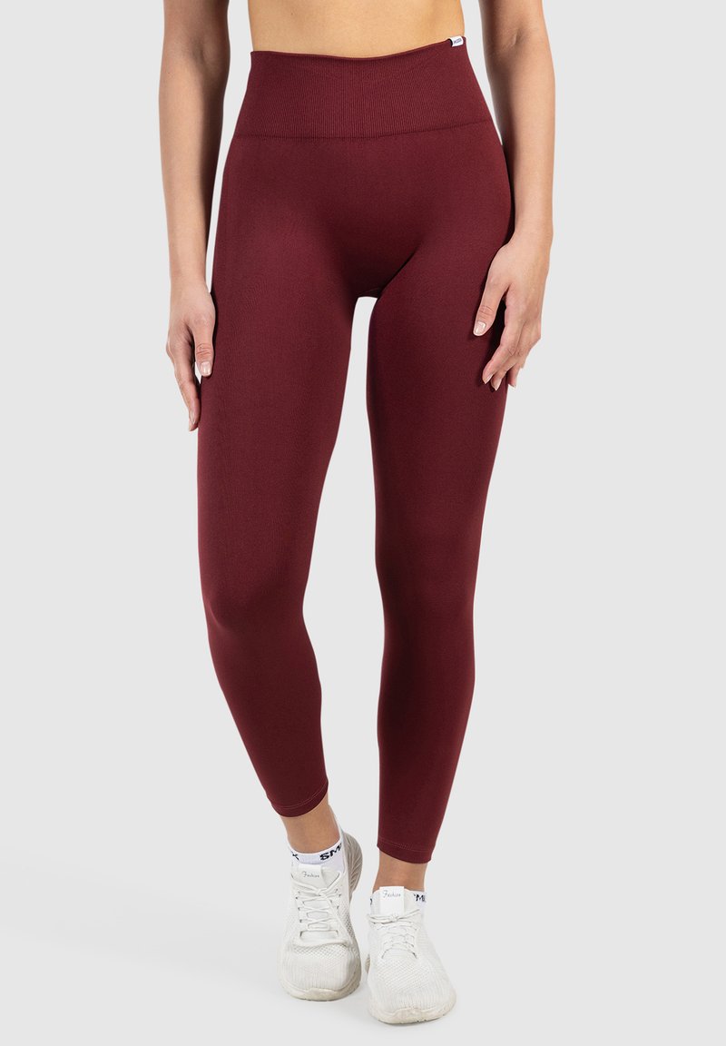 Leggings bordeaux taille haute en tissu extensible, avec une texture lisse et une coupe moulante, associés à des chaussures de sport blanches.