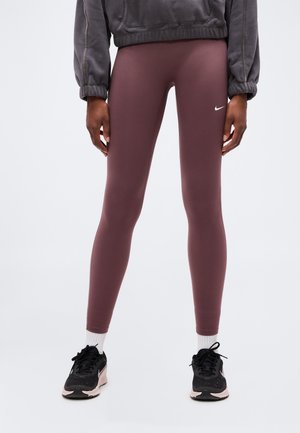 Personne portant des leggings Nike mauves, des baskets noires et roses, des chaussettes blanches et un haut gris à manches longues, debout les mains détendues le long du corps.
