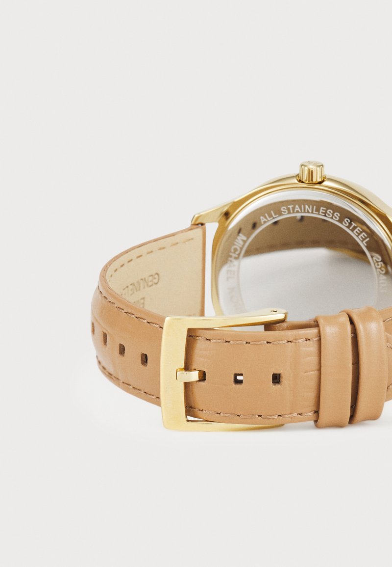 Montre en acier inoxydable doré avec un bracelet en cuir marron clair. Le bracelet présente des coutures, des trous pour le réglage et une boucle en métal poli.