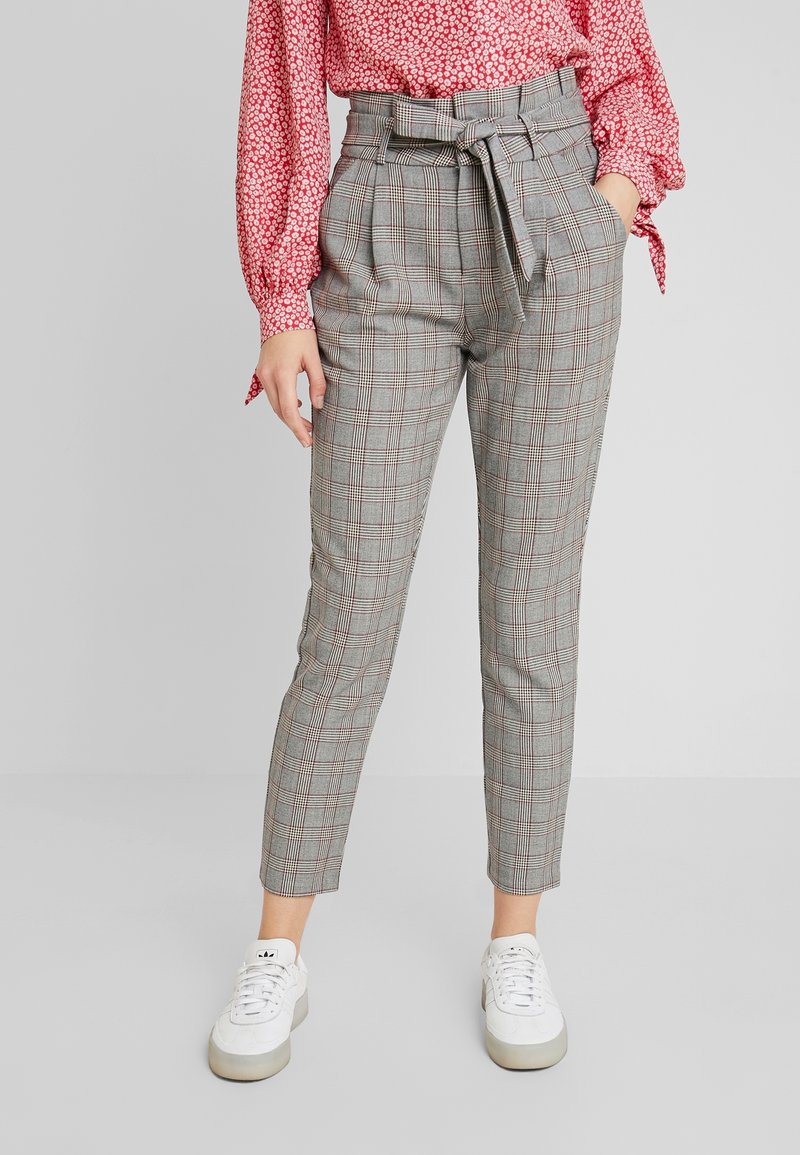Femme portant un pantalon gris à carreaux taille haute avec une ceinture nouée, un chemisier rouge à motifs et des baskets blanches, debout, la main dans la poche.