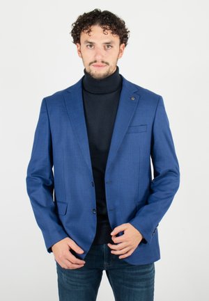 Blazer à carreaux bleus en tissu texturé, avec une fermeture à un bouton, des revers crantés et une poche poitrine. Porté sur un col roulé noir.