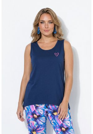 Femme aux cheveux blonds ondulés portant un haut sans manches bleu marine avec un détail cœur rose et un pantalon fleuri coloré sur fond blanc.