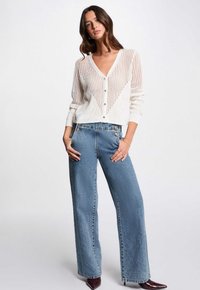 Cardigan bianco lavorato a maglia con motivo a righe verticali e bottoni, abbinato a jeans a vita alta azzurri e a gamba larga e scarpe bordeaux a punta.