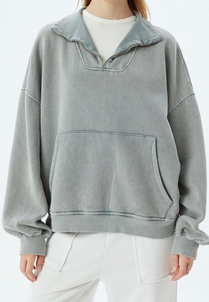 Femme en pull vert délavé oversize avec col demi-fermeture éclair et poche devant, associé à un pantalon blanc ample sur un fond uni.