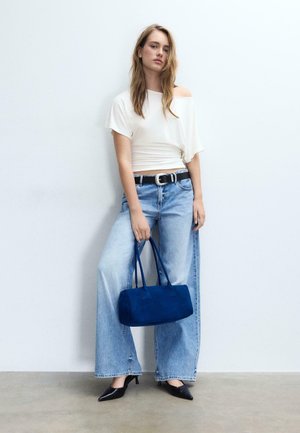 Jeune femme aux cheveux longs portant un top blanc à épaules dénudées, un jean large bleu, des talons noirs, une ceinture noire, et tenant un sac à main bleu, debout contre un mur blanc.