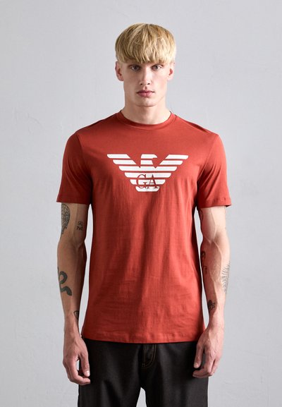Rød bomulds t-shirt med korte ærmer, prydet med et hvidt ørnelogo og stribet design foran, iført af en model med tatoveringer.