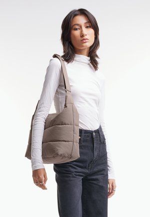 Jeune femme portant un haut blanc à manches longues et un jean noir, avec un grand sac à bandoulière matelassé couleur taupe.