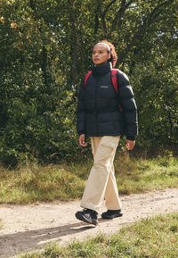 Czarna kurtka puchowa z wysokim kołnierzem i paskiem, beżowe spodnie, czarne buty trekkingowe i czerwona plecak, idąca ścieżką gruntową otoczoną zielenią.