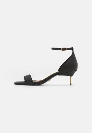 Sandale noire en cuir à bout ouvert avec bride à la cheville, talon stiletto doré bas et semelle plate sur fond blanc.