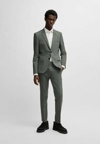 Un costume en tissu vert discret avec un motif subtil ; veste à un bouton et pantalons ajustés. Complété par une chemise blanche et des chaussures noires.