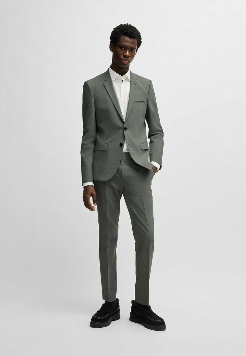 Un costume en tissu vert discret avec un motif subtil ; veste à un bouton et pantalons ajustés. Complété par une chemise blanche et des chaussures noires.