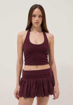 Joven mujer con cabello largo y liso, vestida con un top corto burdeos sin mangas y una falda mini plisada a juego, de pie frente a un fondo liso y claro.