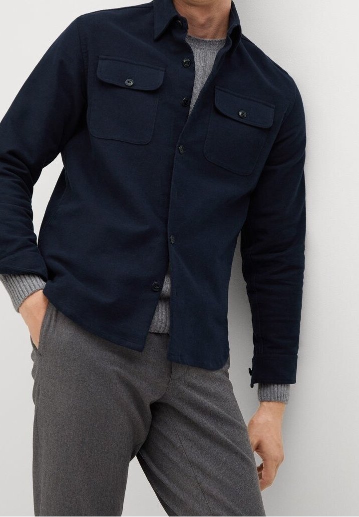 Homme portant une surchemise bleu marine à boutons avec poches à rabat, un pull gris en dessous et un pantalon gris, debout contre un mur uni.