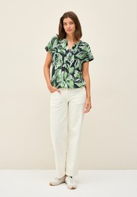 Blusa a maniche corte con un motivo foglia verde e nero, abbinata a pantaloni bianchi a gamba larga e sneaker beige. La blusa ha un design a scollo a V.