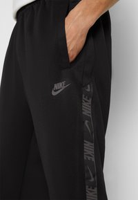 Svarta träningsbyxor med en jämn textur, med en sidostrand som har upprepade svarta Nike-logotyper och en synlig ficka.