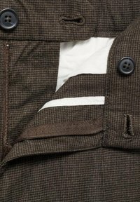 Brauner kariertstoff mit einer Reißverschlusstasche, schwarzen Knöpfen und einem weißen Futterdetail. Enthält ein Knopfloch und eine strukturierte Webart.
